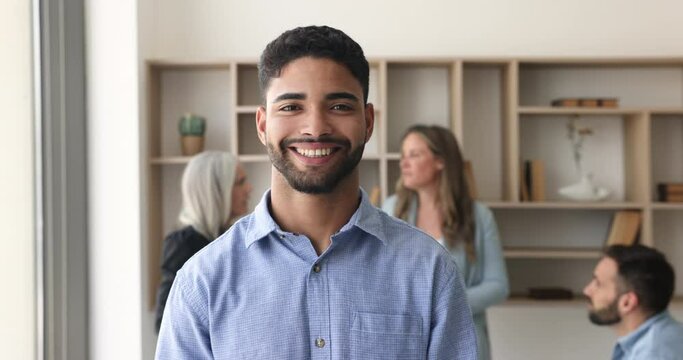 Head shot portrait happy handsome millennial male office employee, newcomer smile look at camera standing in workspace with diverse group of colleagues on background. Career growth, business ambitions