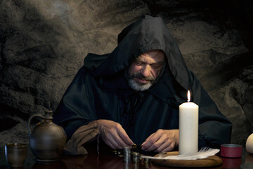 A monk in a robe counts money or coins by candlelight in a dark stone room. A tax collector in...
