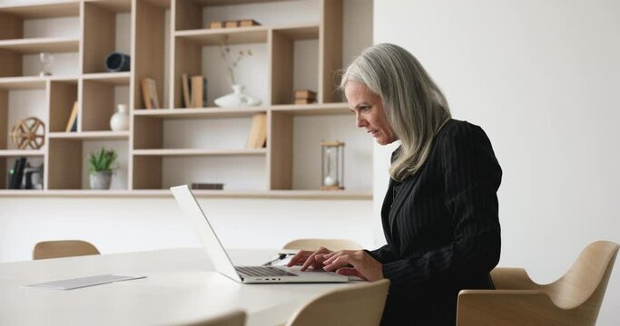 Mature serious businesswoman sits in modern boardroom, working on laptop. Concentrated middle-aged businesslady in suit prepare presentation or financial report for negotiations with corporate clients