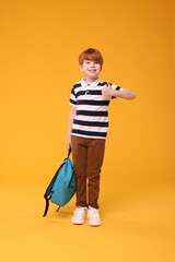 Smiling schoolboy with backpack showing thumb up on orange background