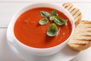 Delicious tomato cream soup in bowl with pieces of grilled bread on white wooden table, closeup