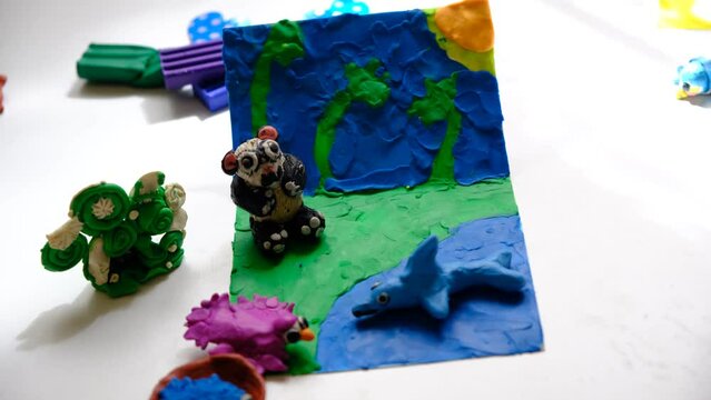 Child smearing colorful plasticine on cardboard and creating fairy tale card with cartoon animals, panda, birds, shark. By spreading, molding and adding texture to plasticine child creating scene