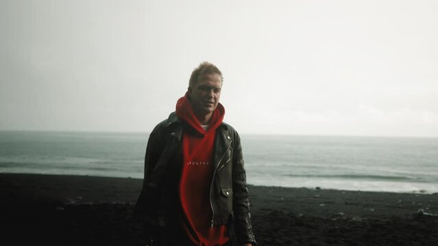 Stylish blonde man in black leather jacket, red hoodie looking at camera, smiling, walking on a rainy day on black-sanded beach with stormy restless sea. High quality 4k footage