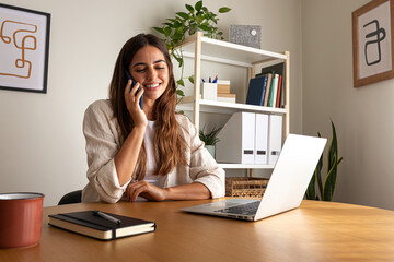 Happy young woman talking on the phone while working at home office using laptop.