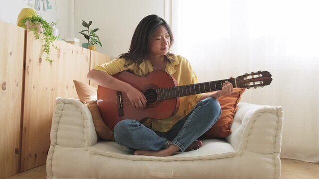Happy teen asian girl composing song sitting on couch. Practicing chords. Playing acoustic guitar. 4k resolution video.