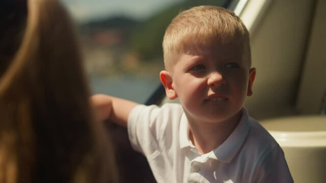 Bored little boy bites lower lip sailing in motorboat salon on sunny day closeup. Funny toddler kid rides vessel along river on summer vacation