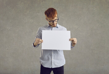 Serious young man holding white blank sheet of paper standing on gray background. Caucasian red-haired man in shirt looking at blank mockup in his hands. Advertising space.