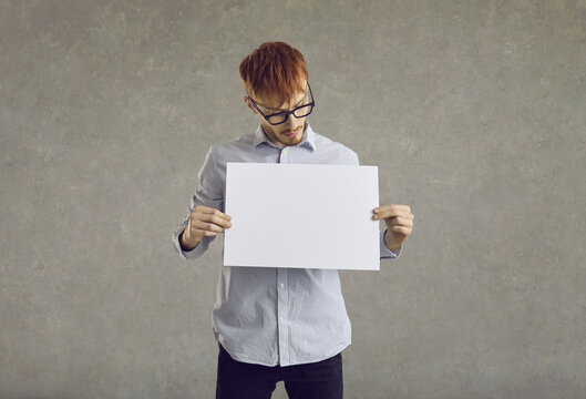 Serious young man holding white blank sheet of paper standing on gray background. Caucasian red-haired man in shirt looking at blank mockup in his hands. Advertising space.