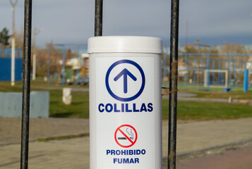 a container to dispose of cigarette butts and a no smoking sign hanging at the entrance to a park, with the words written in Spanish