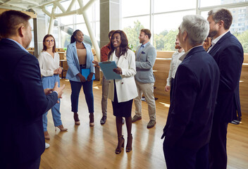 Group of corporate employees meeting with team coach or psychologist. Happy smiling young African American woman with clipboard standing in circle of people during business training event in office