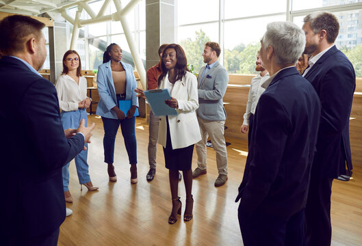 Group Of Corporate Employees Meeting With Team Coach Or Psychologist. Happy Smiling Young African American Woman With Clipboard Standing In Circle Of People During Business Training Event In Office