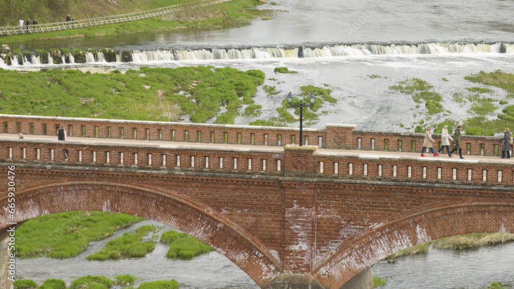 Sticker riga, latvia - august 20 2023. a bridge over a river with a waterfall in the background.