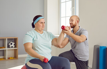 Fat woman doing fitness workout with her trainer for in fitness club. Plus size, obese, overweight woman sitting on fitball and training with dumbbells under guidance of personal trainer