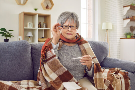 Senior Woman Catches A Cold. Mature Woman Catches The Flu, Has A Fever, Feels Bad And Takes Her Temperature. Sick Old Lady In A Warm Plaid And Glasses Sitting On The Sofa At Home Holding A Thermometer