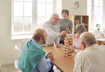 Cheerful retired friends playing jenga board game at home. Happy senior men and women sitting...
