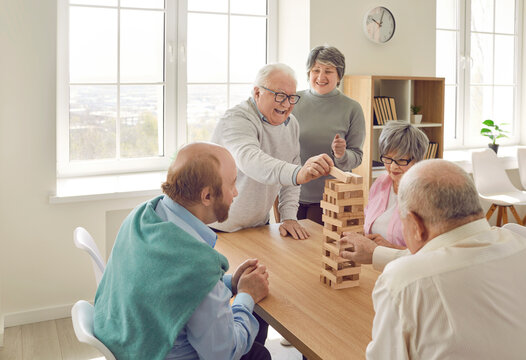 Cheerful retired friends playing jenga board game at home. Happy senior men and women sitting around table playing and communicating in living room. Elderly people leisure activities, nursing home