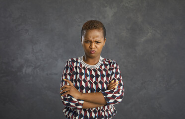 Portrait of displeased grumpy mad young black woman pouting lips and looking at camera with expression of displeasure and resentment on face standing arms folded crossed against grey studio background