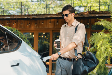 Eco friendly man and sustainable urban commute with EV electric car recharging at outdoor coffee...