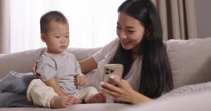 Asian mom and toddler spending time together. Multiethnic asian family, mom and son, woman and boy, mommy mother showing toddler baby video on smartphone. Boy watching cartoons with mom using phone.