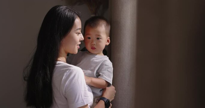 Asian mom and baby boy together. Multinational mother holds child in arms looks dreamily thoughtfully out of window smiles gently lovingly hugs son kisses on cheek. Mom and kid at window. Slow motion