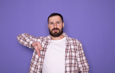 Portrait of unsatisfied bearded man with thumbs down against purple background. Studio shot.