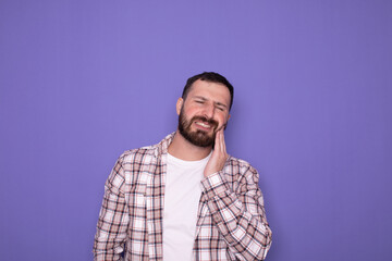 Dental problems. Portrait of unhealthy man pressing sore cheek, suffering acute toothache, periodontal disease, cavities or jaw pain. Indoor studio shot isolated on purple background. 