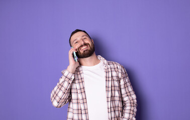 Young smiling happy bearded caucasian man speaking by mobile cell phone spread hand isolated on purple background studio portrait. People lifestyle concept.