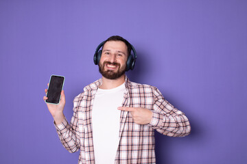 Portrait bearded smiling young man in wireless headphones pointing at smartphone on purple background. Millennial guy promoting new mobile app, listening to music. Lifestyle. 