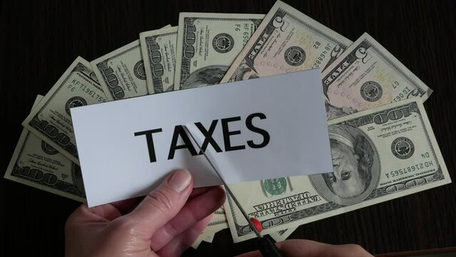 the cutting paper with inscription taxes lies on top of a large pile of dollar USA money on a dark wooden table, top view, tax reduction