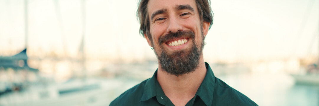 Close-up portrait of a smiling man with a beard on the embankment, on a yacht background. Frontal closeup of happy young hipster male looking at camera