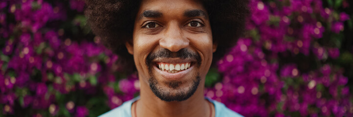Close-up frontal portrait of young African American man opens his eyes looks at the camera and smiling standing on background wall covered with purple flowers. Urban lifestile
