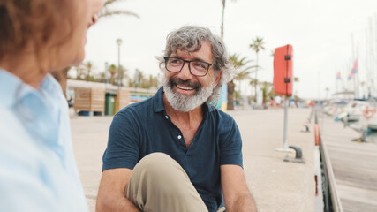 Beautiful middle aged woman sitting in the port and talking with her husband on yacht background