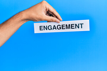 Hand of caucasian man holding paper with engagement word over isolated blue background