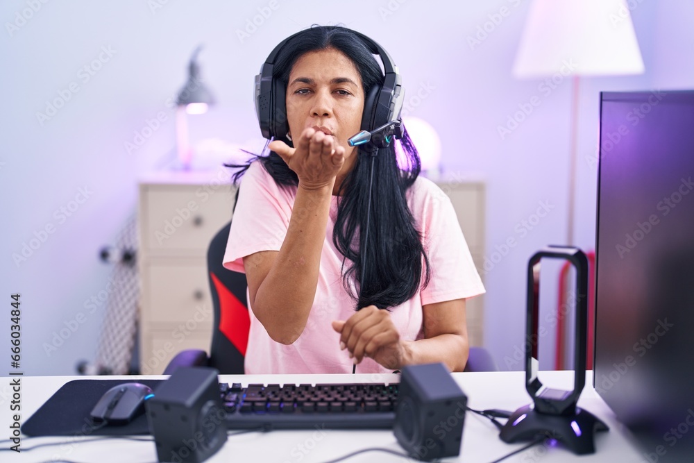 Poster mature hispanic woman playing video games at home looking at the camera blowing a kiss with hand on 