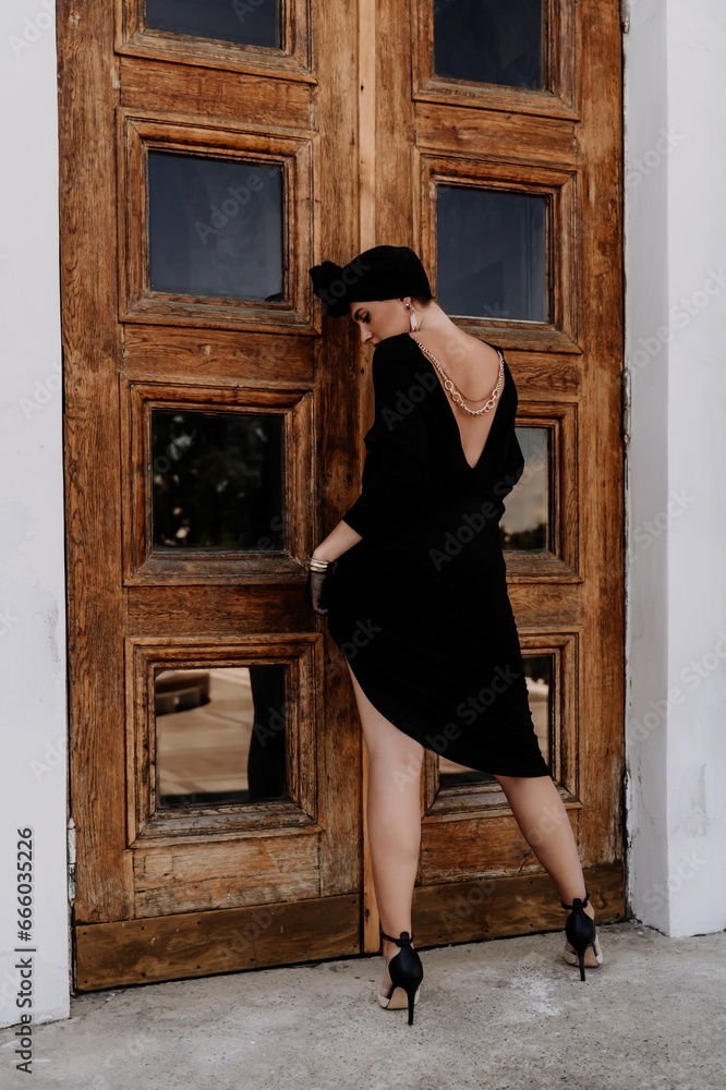 Wall mural Stylish woman in the city. Fashion photo of a beautiful model in an elegant black dress posing against the backdrop of a building on a city street