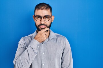 Middle east man with beard standing over blue background looking confident at the camera with smile with crossed arms and hand raised on chin. thinking positive.