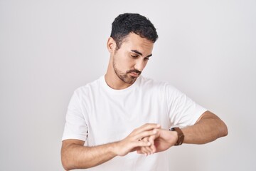 Handsome hispanic man standing over white background checking the time on wrist watch, relaxed and confident