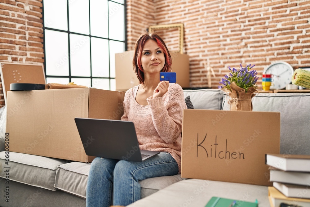 Poster Young caucasian woman using laptop and credit card sitting on sofa at new home