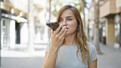 Stunning young blonde woman standing relaxed on city street, engrossed in sending intimate voice...