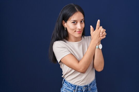 Young hispanic woman standing over blue background holding symbolic gun with hand gesture, playing killing shooting weapons, angry face