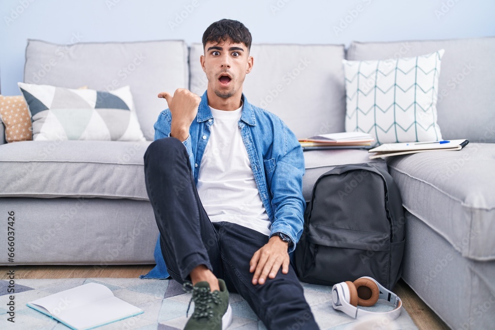 Wall mural young hispanic man sitting on the floor studying for university surprised pointing with hand finger 