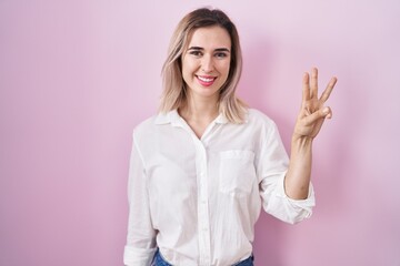 Young beautiful woman standing over pink background showing and pointing up with fingers number three while smiling confident and happy.