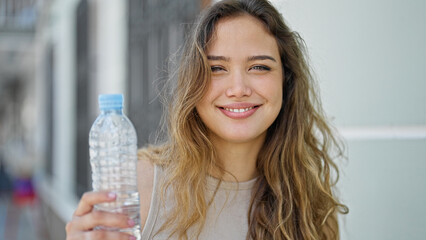 Young beautiful hispanic woman holding bottle of water smiling at street