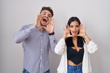 Young hispanic couple standing over white background smiling cheerful playing peek a boo with hands showing face. surprised and exited