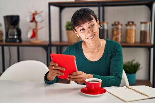 Young beautiful hispanic woman using touchpad drinking coffee sitting on table at home