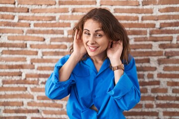 Beautiful brunette woman standing over bricks wall trying to hear both hands on ear gesture, curious for gossip. hearing problem, deaf