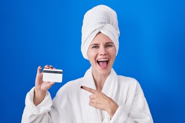 Blonde caucasian woman wearing bathrobe holding credit card smiling happy pointing with hand and finger