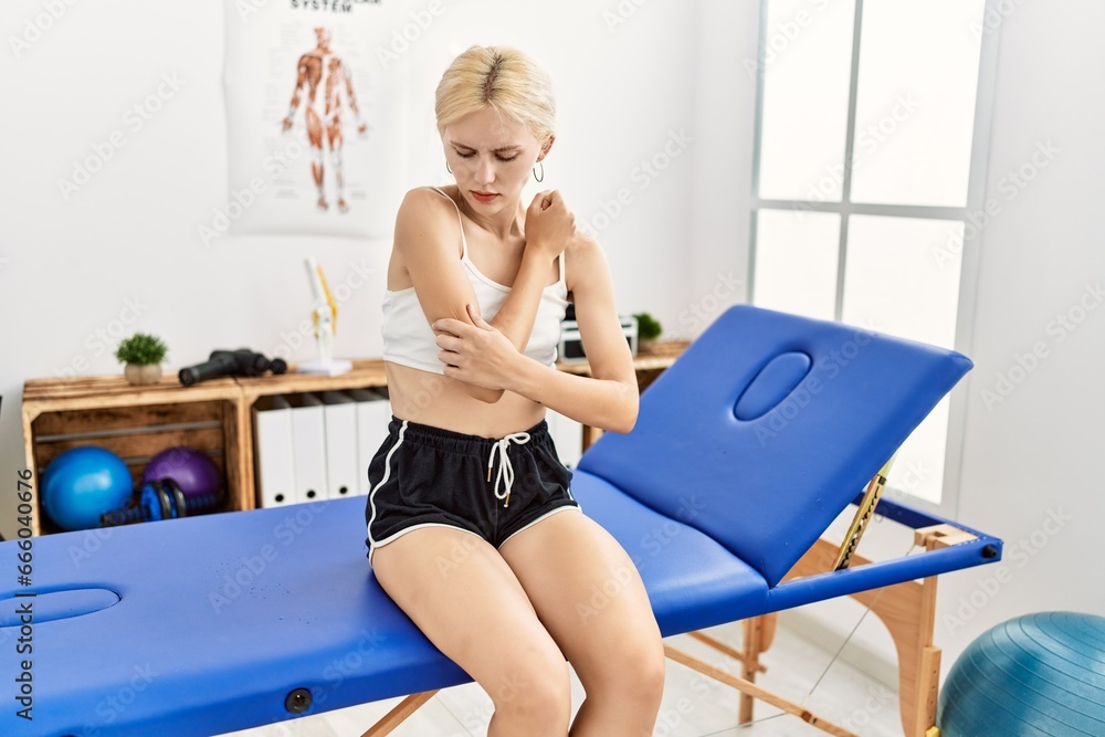 Poster young blonde woman patient suffering for arm pain sitting on massage table at rehab clinic