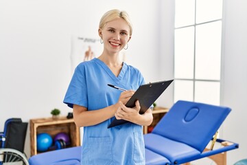 Young blonde woman pysiotherapist smiling confident writing on document at rehab clinic