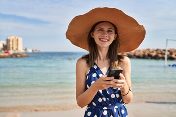 Fototapeta premium Young blonde woman tourist wearing summer hat using smartphone at beach